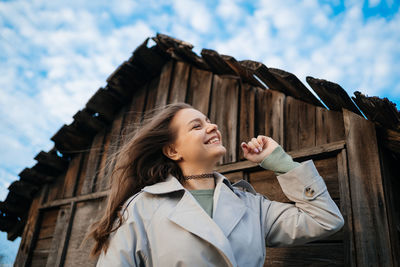 Beautiful girl with long hair in a grey trench coat next to an old wooden house blue sky background 