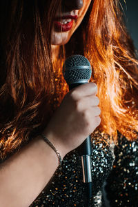 Faceless portrait of redhead woman in sparkly evening dress holding microphone on dark night