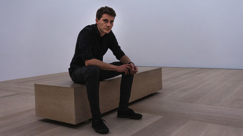 Young man looking away while sitting on hardwood floor against wall