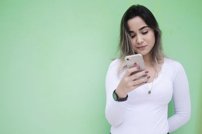 Young woman using phone while standing against wall