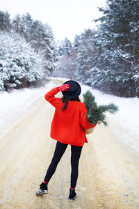 A girl in a red sweater and hat stands in the middle of a snowy road in a forest with pine branches