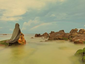 A woman wearing hijab sitting on rock by the sea against sky looking at rainbow