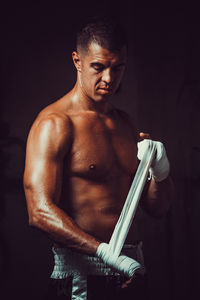 Shirtless male athlete wearing hand wrap while standing in darkroom