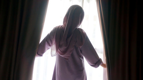 Rear view of woman standing by window at home