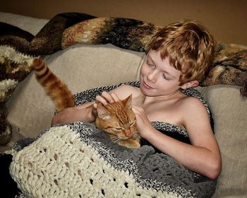 High angle view of shirtless boy playing with cat in knitted blanket