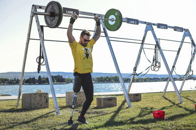 Male adaptive athlete lifting deadlift while standing against lake at park during sunny day