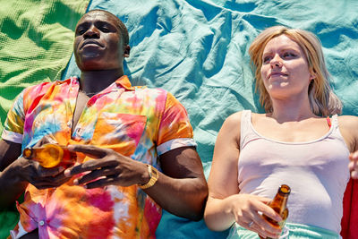 Top view of relaxed young multiracial couple in casual clothes lying on blanket with bottles of beer and looking away during summer holidays