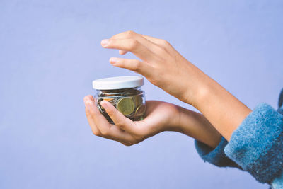 Cropped hand of woman holding coins in glass container against purple background