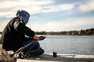 Man fishing while sitting on pier over lake against sky