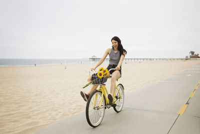 Cheerful woman cycling on street by beach against clear sky