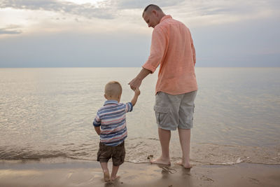 Rear view of father holding son's hand while standing on shore at beach