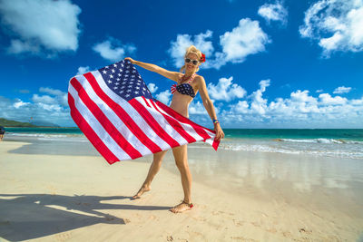 Portrait of happy woman holding american flag while standing at beach against blue sky