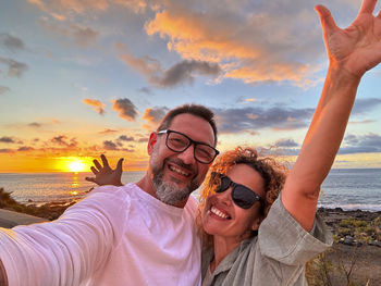 Happy friends with arms raised standing at beach against sky during sunset