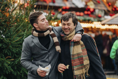 Smiling man eating trdelnik with arm around father enjoying at christmas market