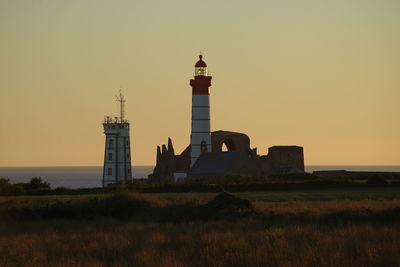 Lighthouse on field by building against sky during sunset