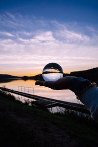 Cropped hand of woman holding crystal ball against sky during sunset
