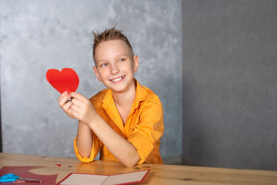 A cute boy is sitting at a table and making valentine's day cards with red hearts