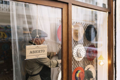 Through glass side view of concentrated mature male seller hanging stylish assorted hats behind showcase in fashion boutique