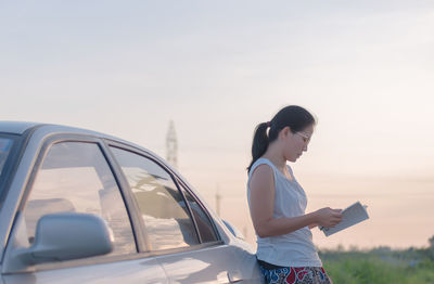 Beautiful young woman reading book by car on road