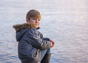 Portrait of boy wearing warm clothing sitting on rock by lake during sunset