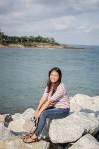 Portrait of young woman sitting on rock by sea against sky