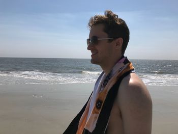 Side view of shirtless young man with towel standing at beach against sky during sunny day