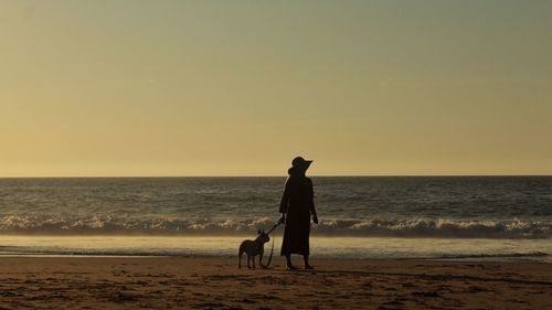 Woman with dog standing on shore at beach against sky during sunset