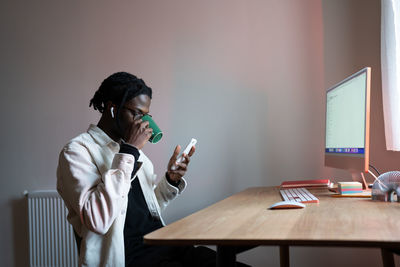 Relaxed carefree man drinking tea sits at desk and using mobile phone sits at table with computer