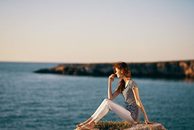 Woman sitting by sea against sky during sunset