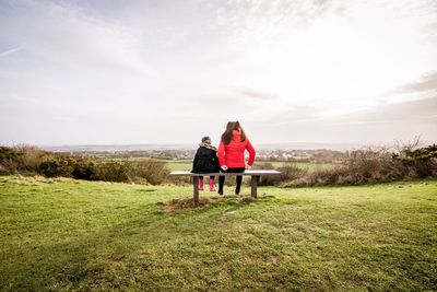 Rear view of mother and daughter sitting on bench over field against sky