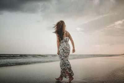 Rear view of woman walking at beach against sky during sunset