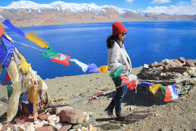Full length side view of woman wearing warm clothing while standing by prayer flags and lake