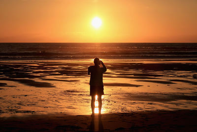 Rear view silhouette of single woman takong photo of sunset standing on a beach.
