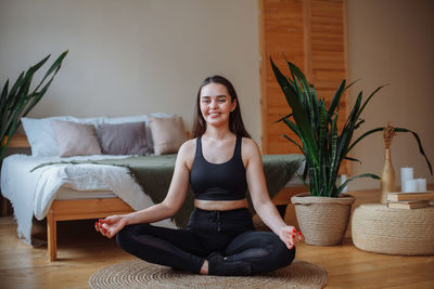 Smiling young woman sits at home on a jute rug near the bed in the lotus position. does yoga at home