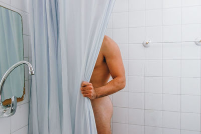 Midsection of naked man standing behind curtain in bathroom at home