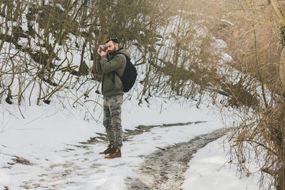 Young man photographing on snow covered road at forest during winter