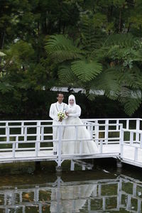 Portrait of newlywed couple standing on pier over lake against trees