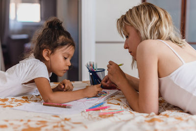 Caucasian mom and hispanic daughter painting while they are lying in the bed.