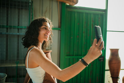 Smiling young woman taking selfie with mobile phone while standing against wall