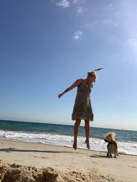 Low angle view of woman jumping by dog at beach against sky