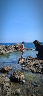 Full length of shirtless man on rock in sea against sky