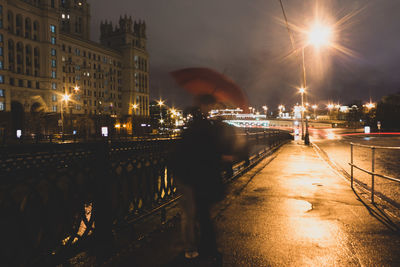 Illuminated street by buildings against sky at night