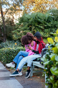 Young black woman sitting on bench and demonstrating smartphone screen to attentive sister while spending time in green park