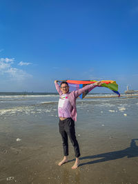 Portrait of a young man standing with a rainbow flag on the beach against the sky