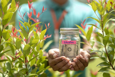Midsection of man holding currency in jar with education text by plants