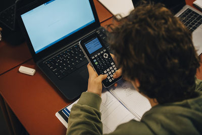 High angle view of teenage boy using calculator while studying over laptop at desk in classroom
