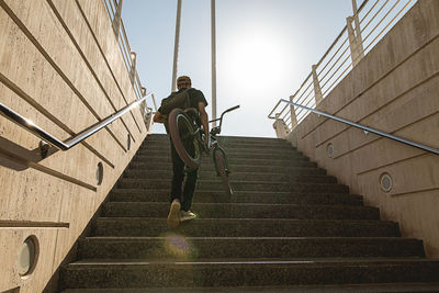 Low angle view of man moving up with bicycle on staircase against sky
