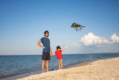 Father and son are standing on a sandy beach by the sea and launch a toy striped kite in the summer