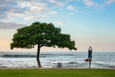 Rear view of woman doing yoga at beach against sky during sunset