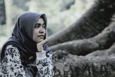 Young woman wearing hijab looking away sitting by tree trunk